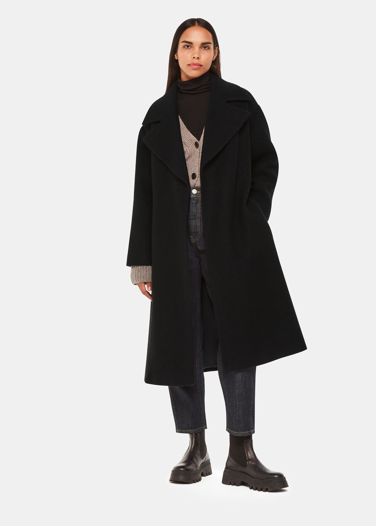 A woman wearing a long black wool coat with a tie waist belt, over a black turtleneck and jeans, paired with black chunky boots.