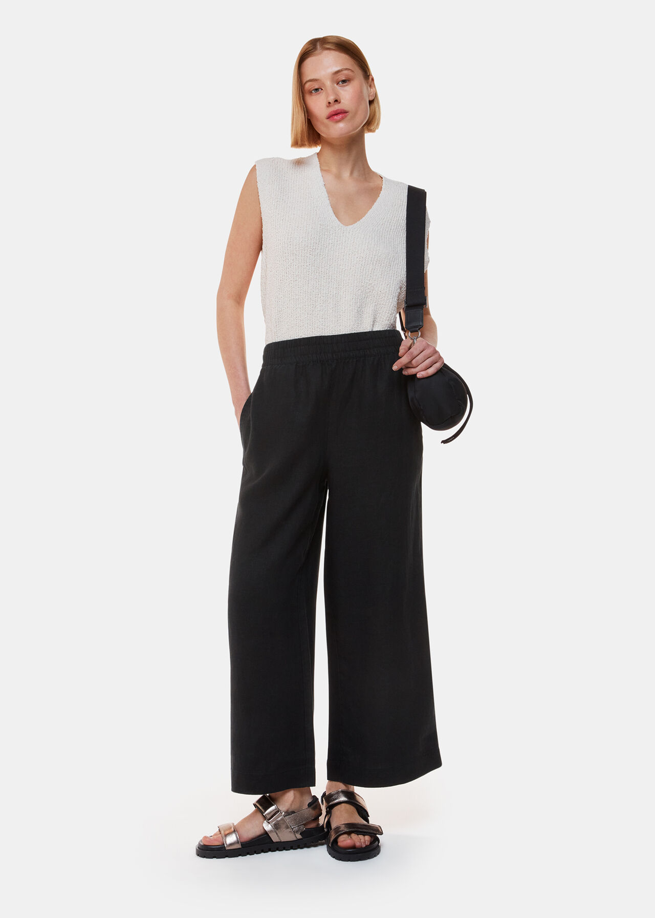 A woman wearing black linen trousers with an elastic waistband and side pockets, paired with a sleeveless white top and sandals.