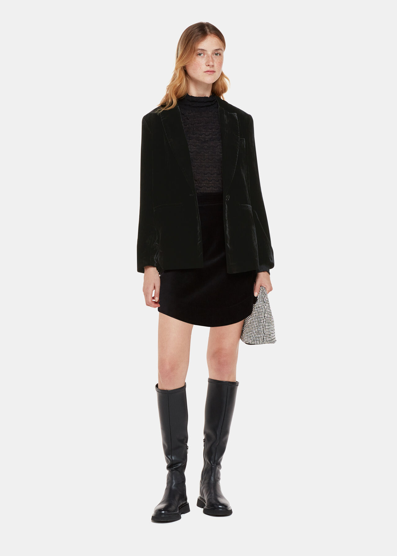 A woman wearing a black velvet blazer with a button closure, long sleeves, and a classic fit, paired with a black skirt and knee-high black boots.