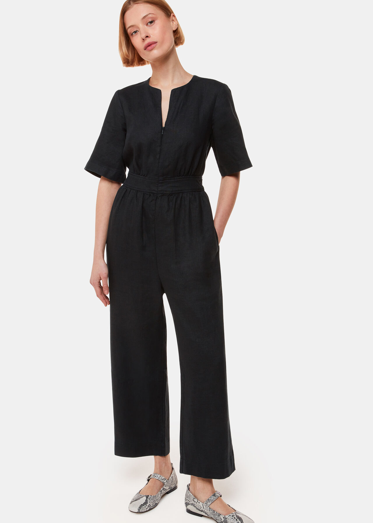 A woman wearing a black linen jumpsuit with short sleeves, a V-neckline, and wide-leg pants, standing against a plain background.