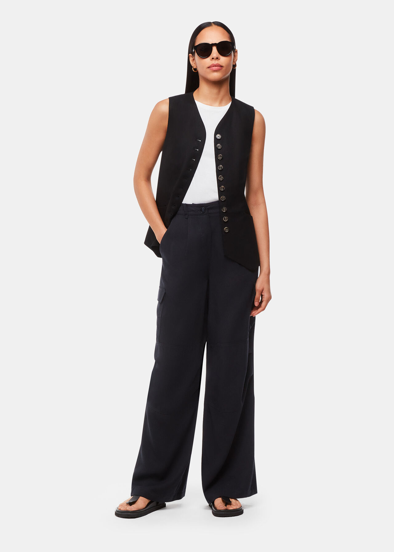 A woman wearing navy blue Luxe Cargo Pants, a sleeveless black vest, white top, black sandals, and sunglasses.
