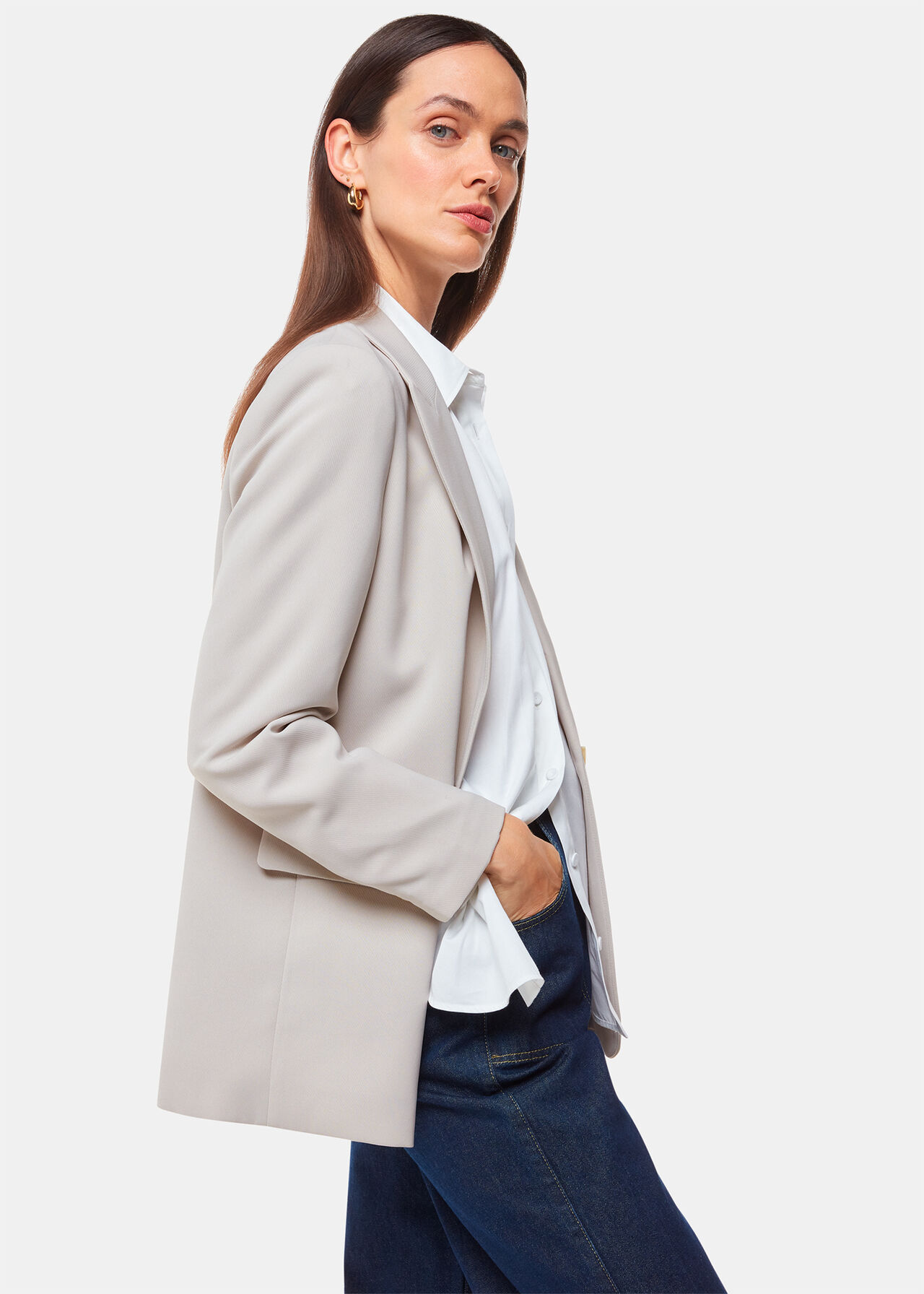 A woman wearing a beige crepe blazer with peak lapels, a button closure, and a relaxed, boxy fit. She has long brown hair and is dressed in a white shirt and dark jeans.