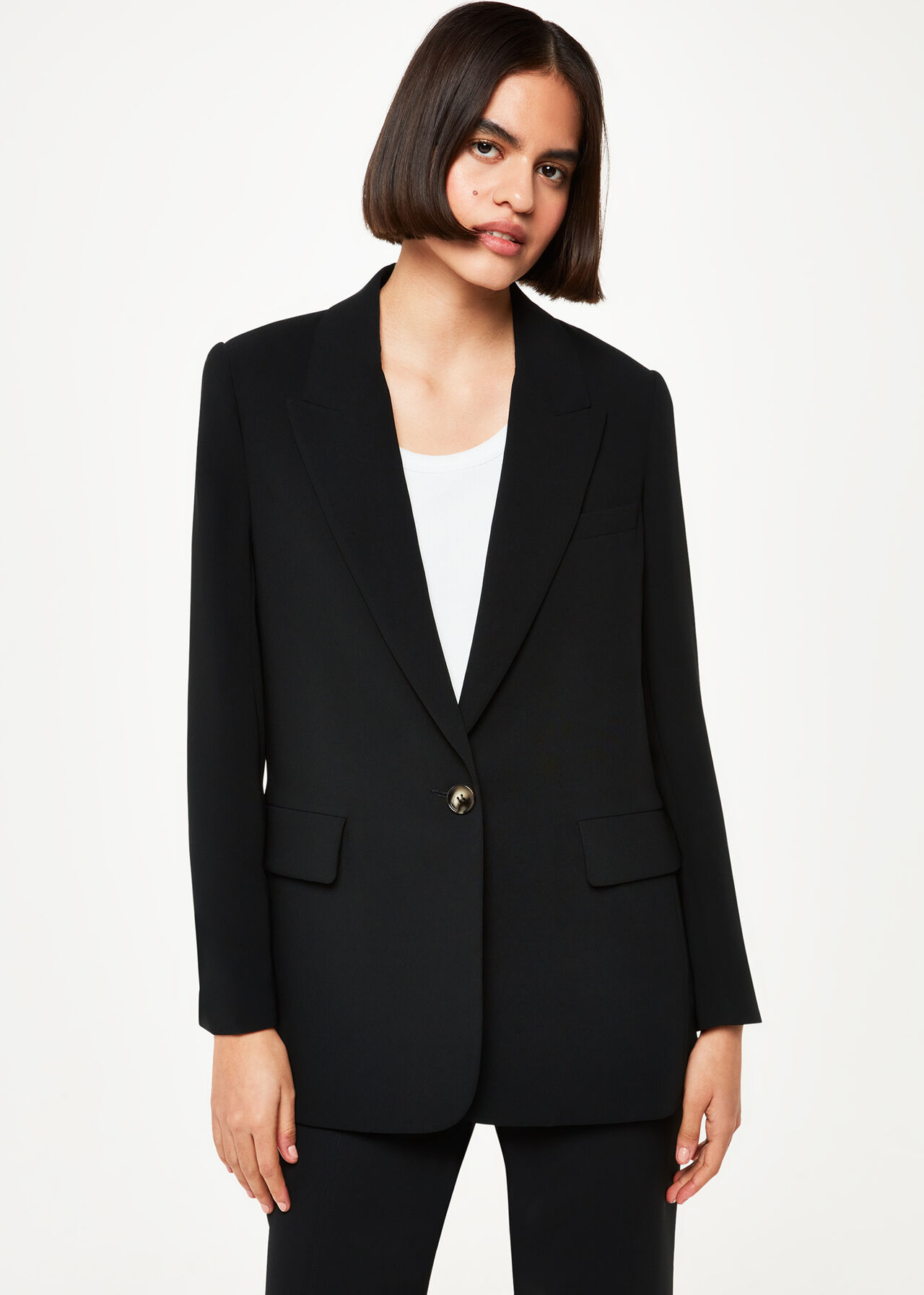 A woman wearing a black crepe boyfriend blazer with peak lapels, relaxed fit, and flap pockets, paired with black pants.