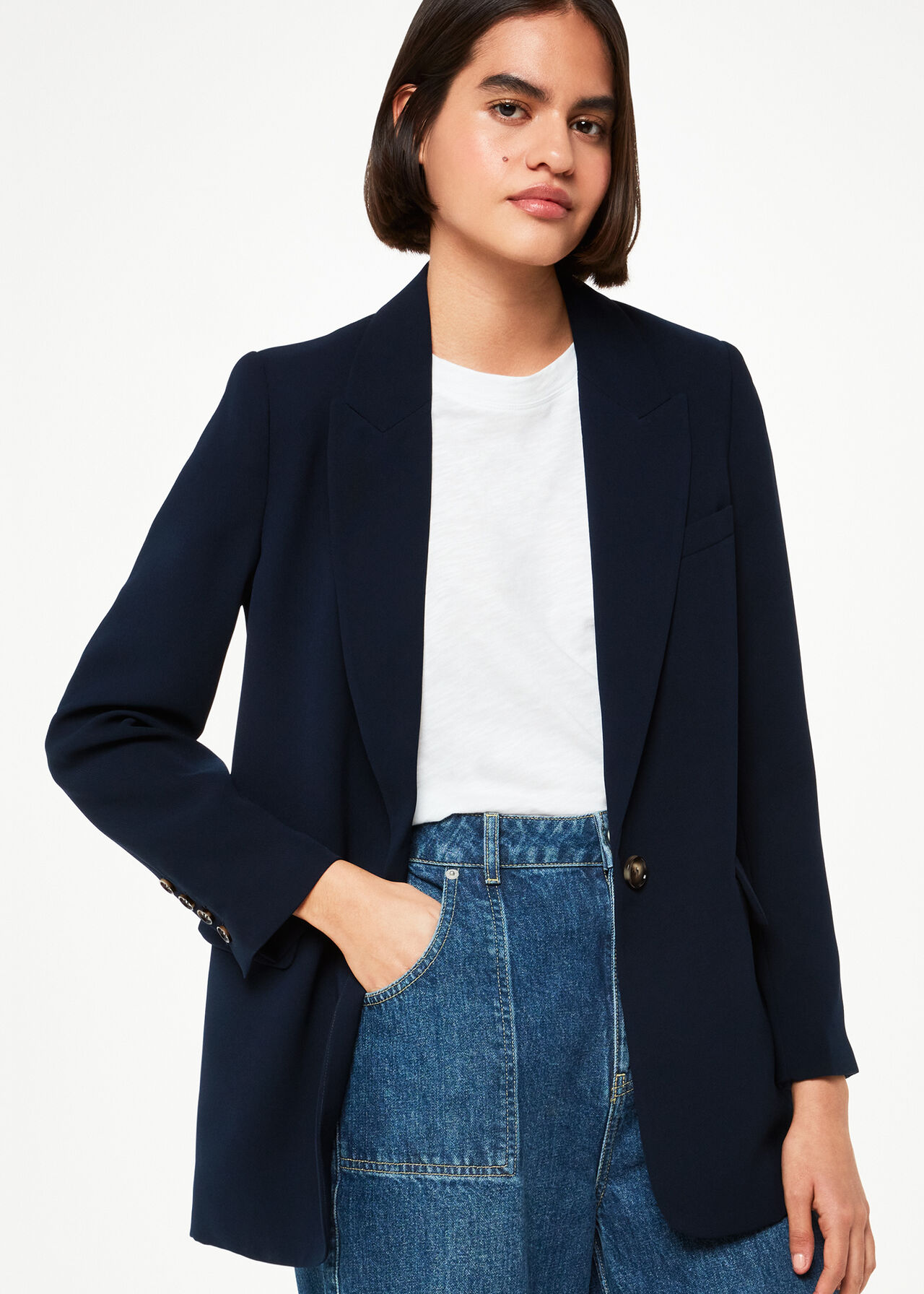 A woman wearing a navy crepe boyfriend blazer with peak lapels, a collared neck, and two side pockets, paired with a white t-shirt and blue jeans.