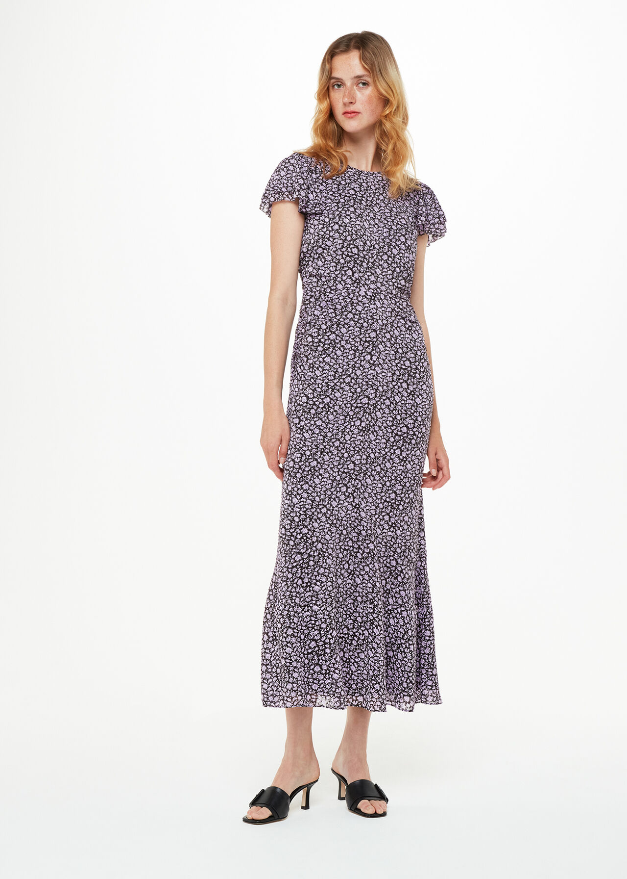 A woman wearing a purple dress with a daisy print, short sleeves, and a gathered waist, standing against a plain white background.