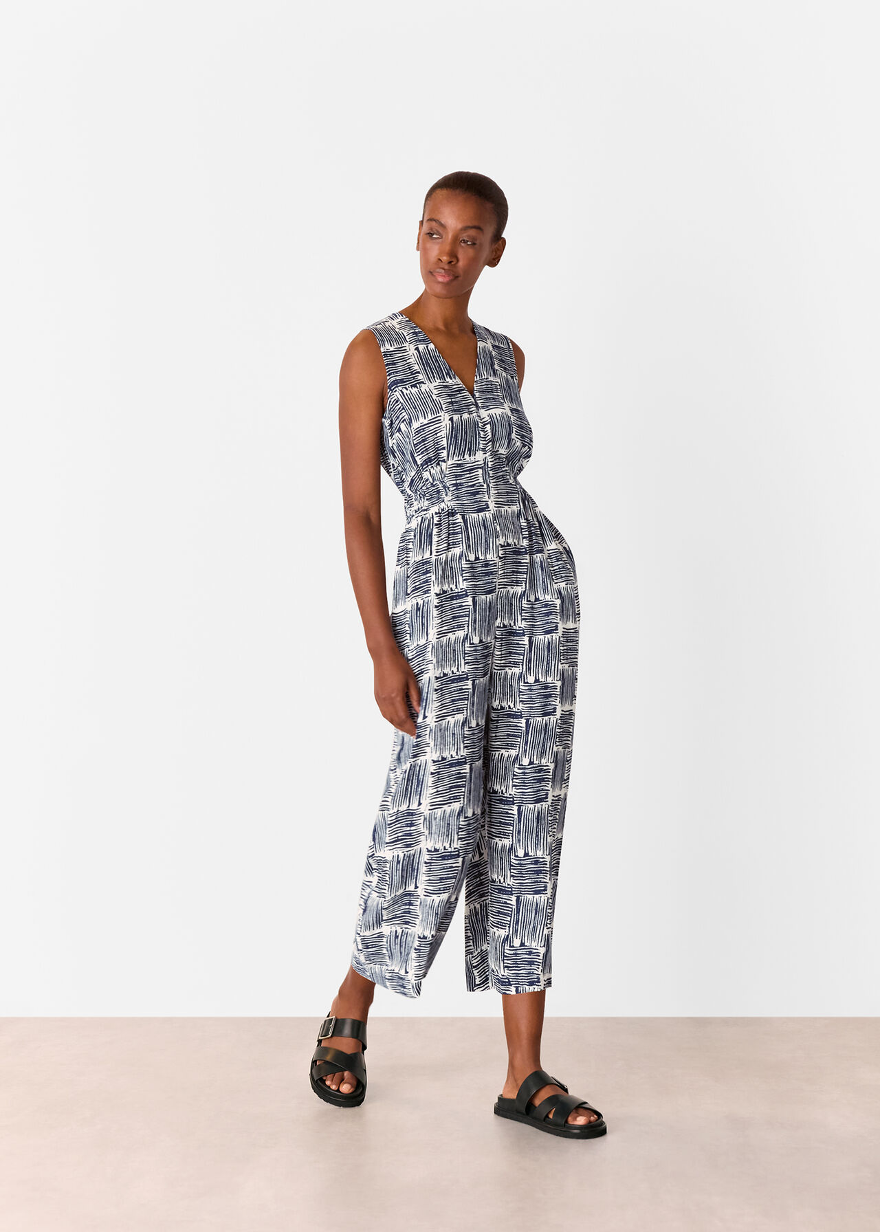 A woman wearing a navy hatch squares jumpsuit with a sleeveless, V-neck design, and cropped straight legs.