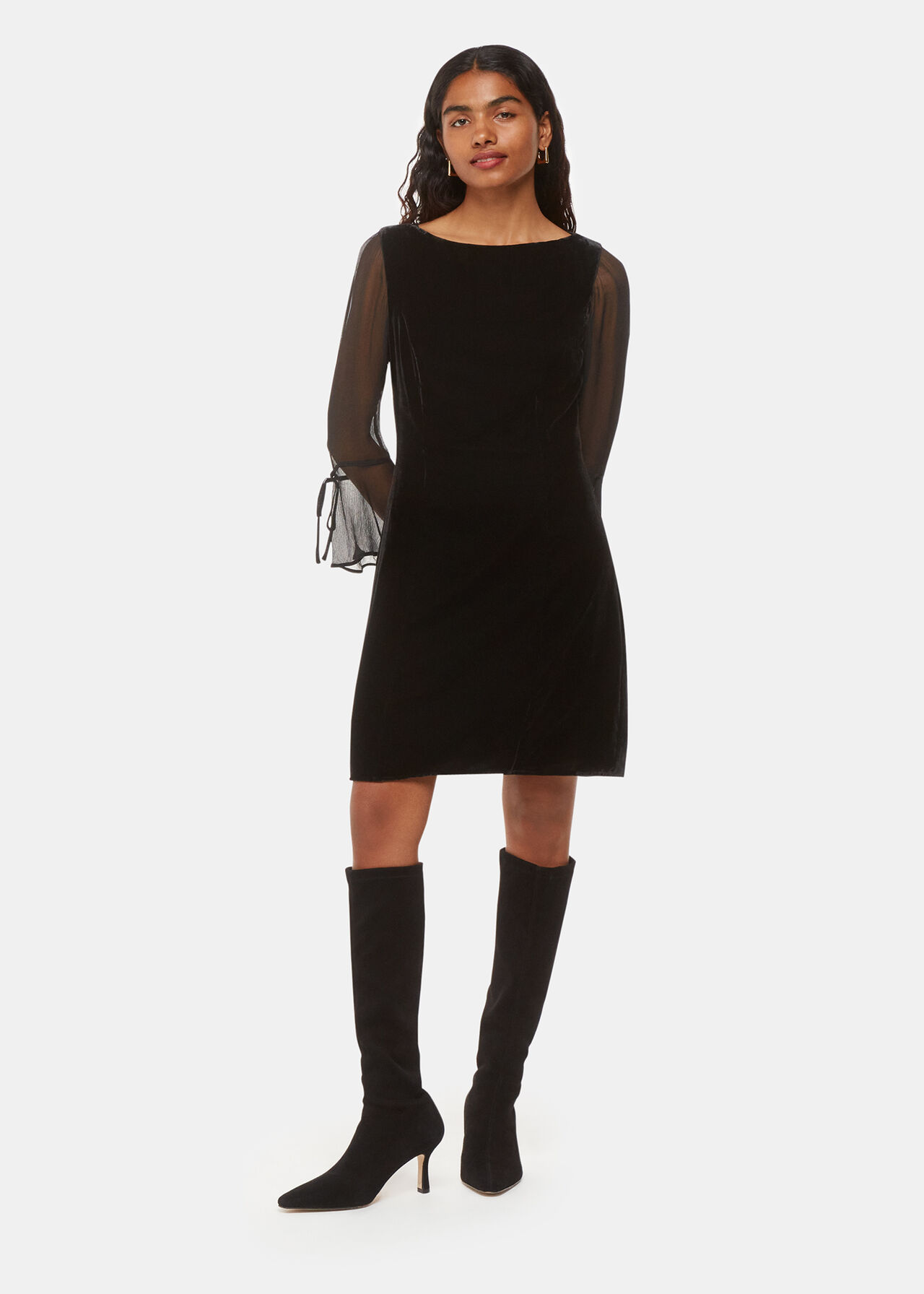 A woman wearing a black velvet mini dress with long mesh sleeves and tie detail cuffs.