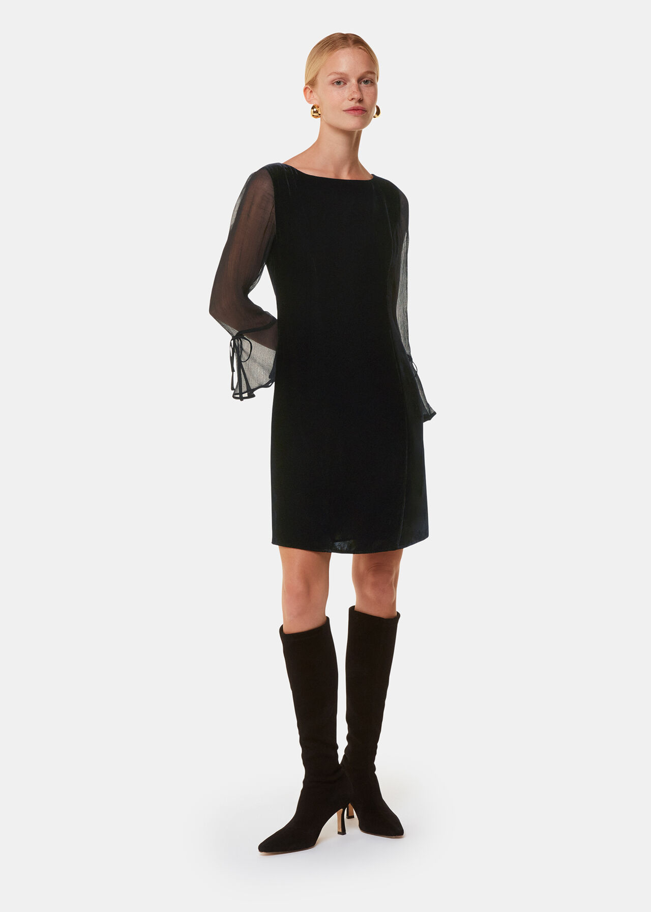 A woman wearing a black velvet mini dress with long mesh sleeves and tie detail cuffs, standing against a plain background.