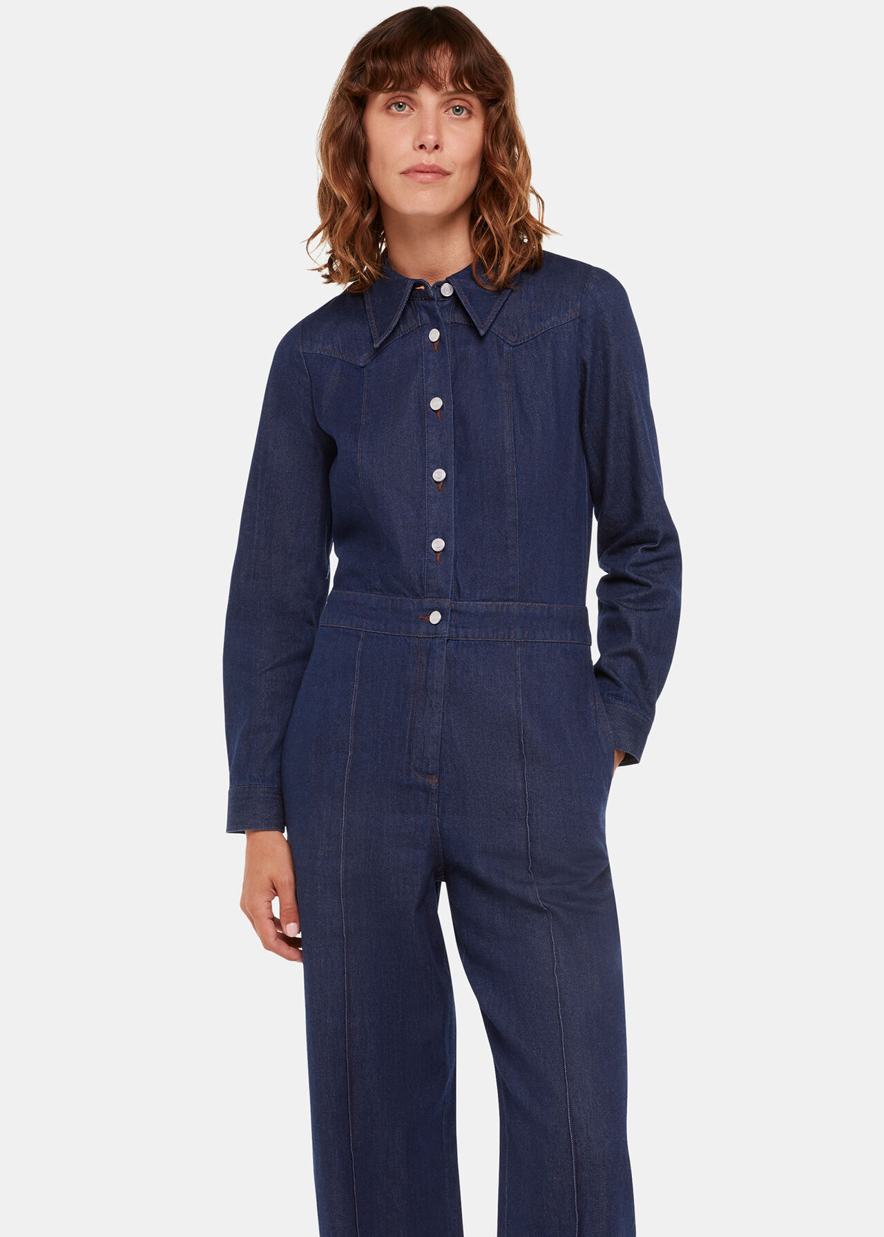 A woman wearing a dark blue denim jumpsuit with a collar, long sleeves, and button-down front, standing against a plain background.