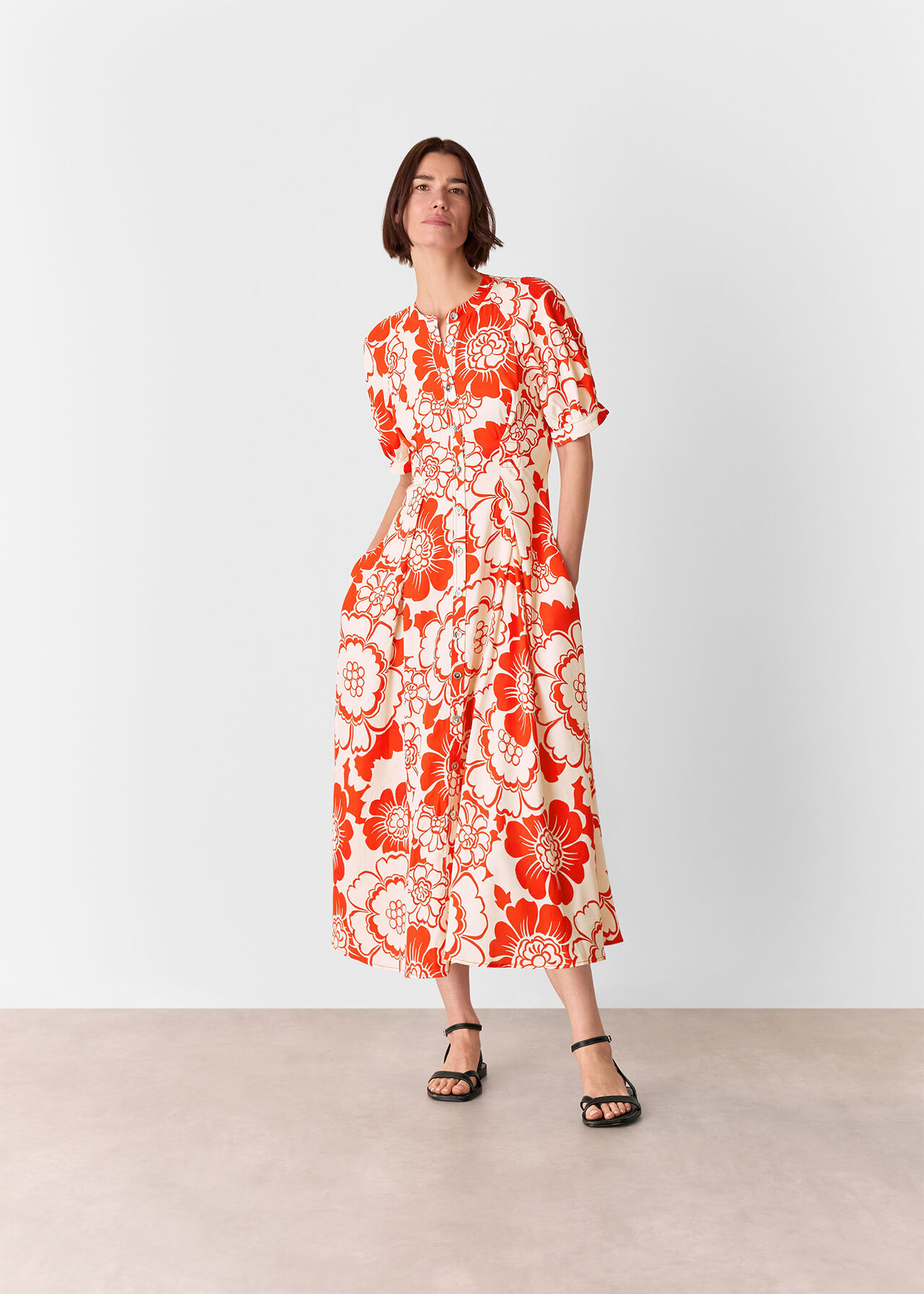 A woman wearing a long, floral midi dress with red and cream colors, short sleeves, and a round neckline, standing against a plain background.