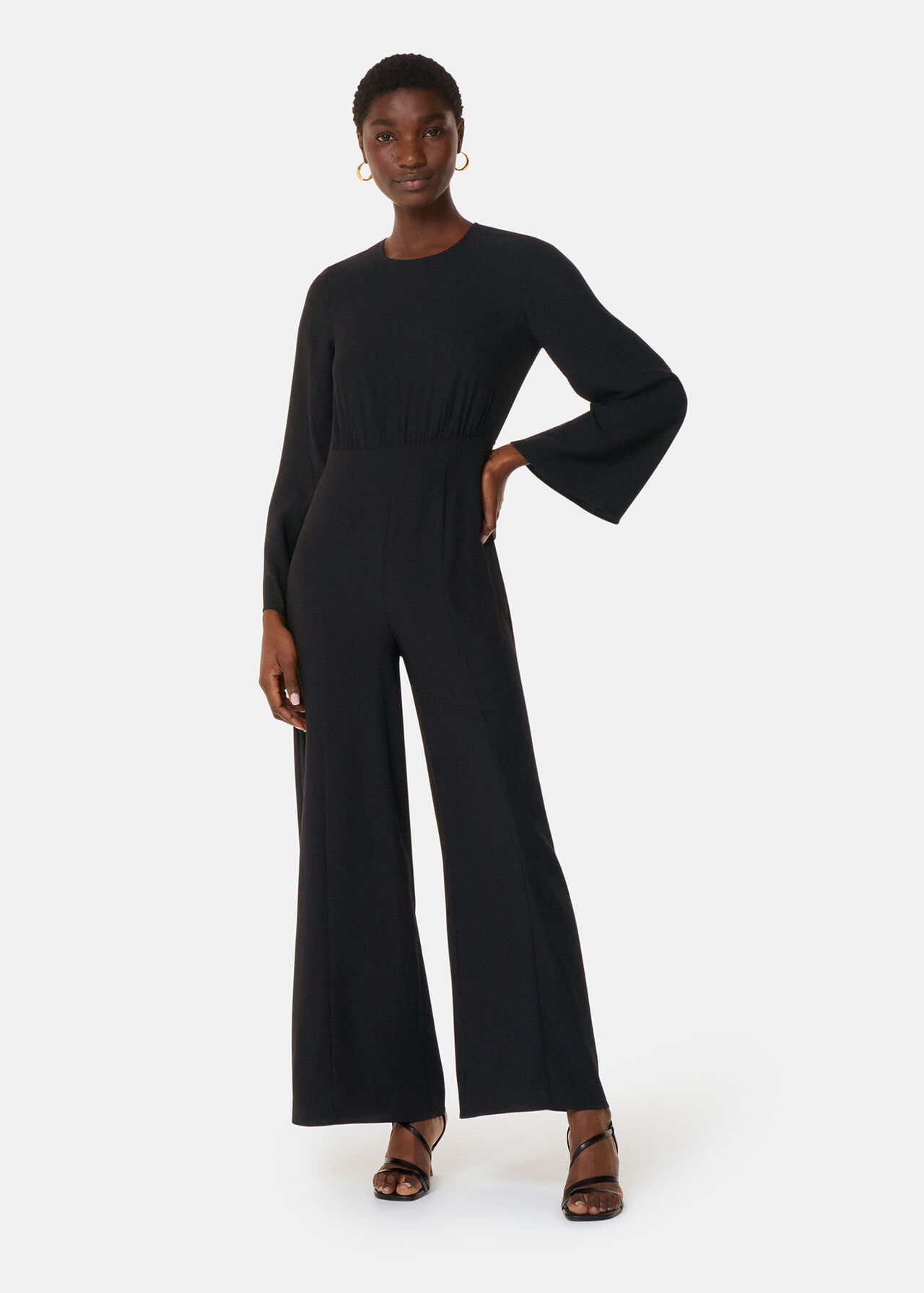 A woman wearing a black gathered jumpsuit with long sleeves, round neckline, and a cinched waist, standing against a plain background.