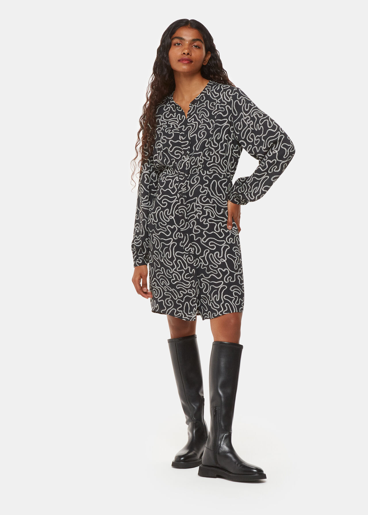 A woman wearing a black dress with a white chain print, long sleeves, and black knee-high boots.