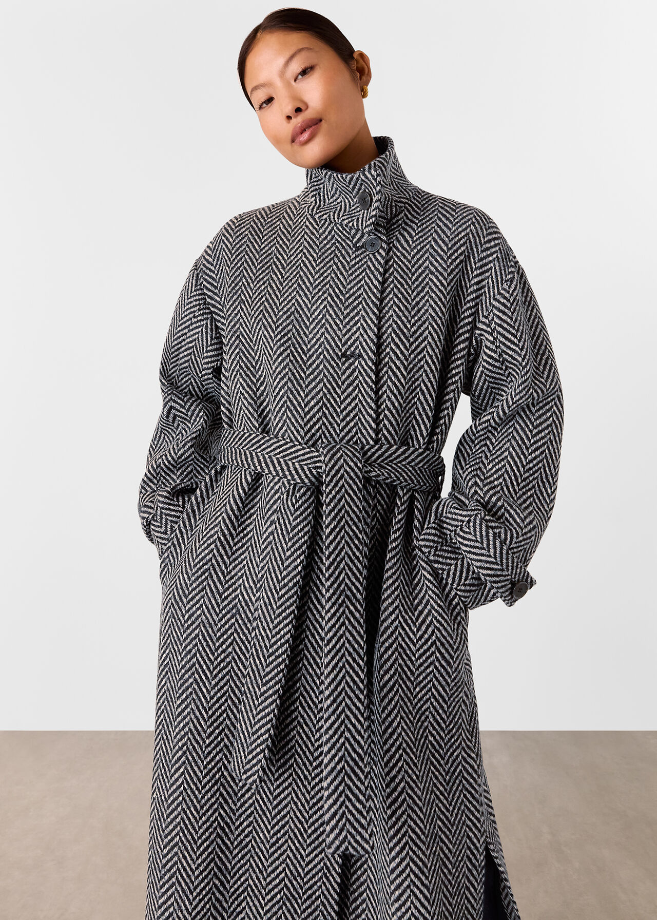 A woman wearing a multicolour Petite Cosette funnel neck coat with a high collar, wrap style, button closure, and a belt at the waist, featuring a textured pattern in black, white, and grey.
