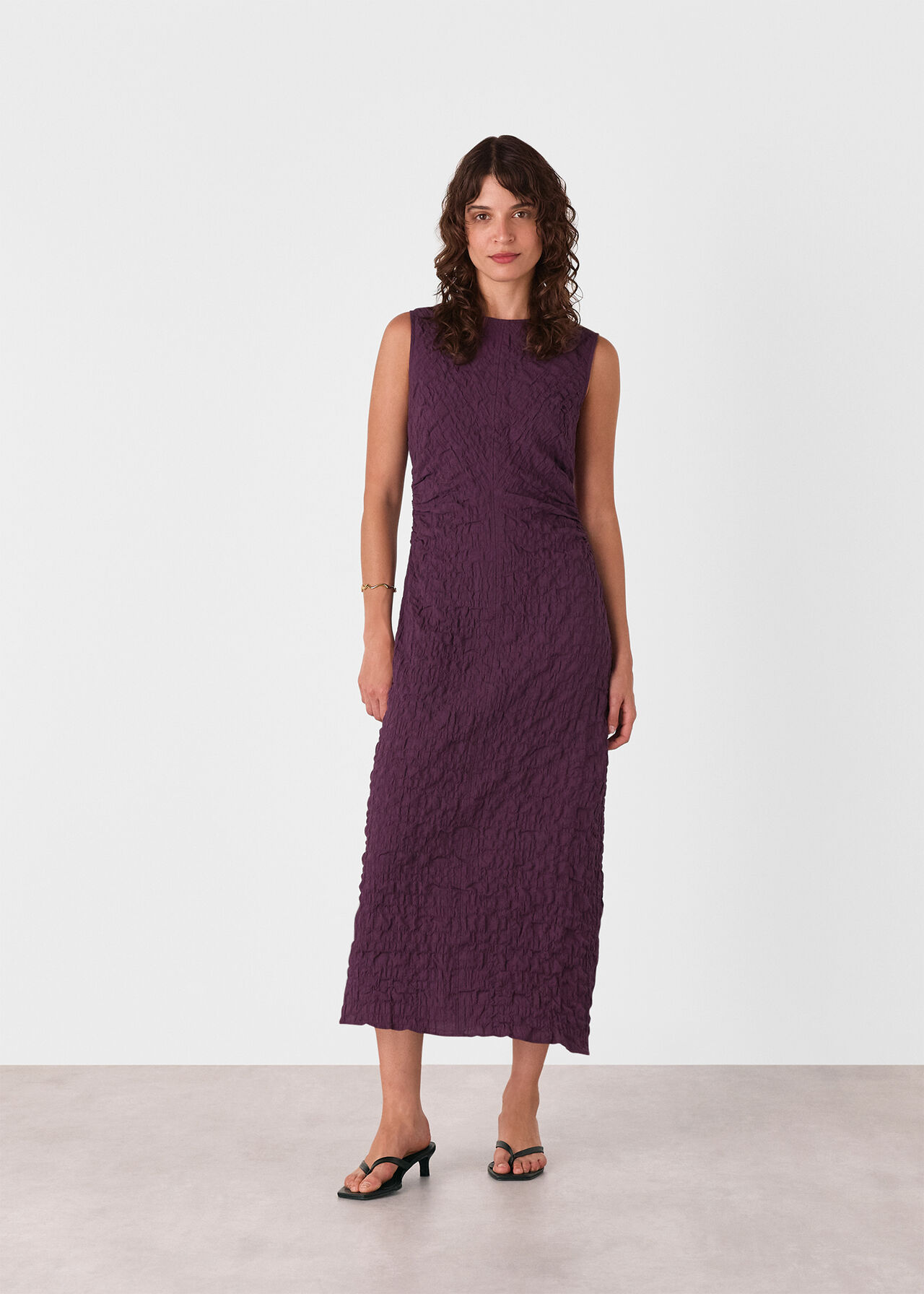 A woman wearing a sleeveless, textured plum midi dress with a round neckline and ruched details, standing against a plain background.