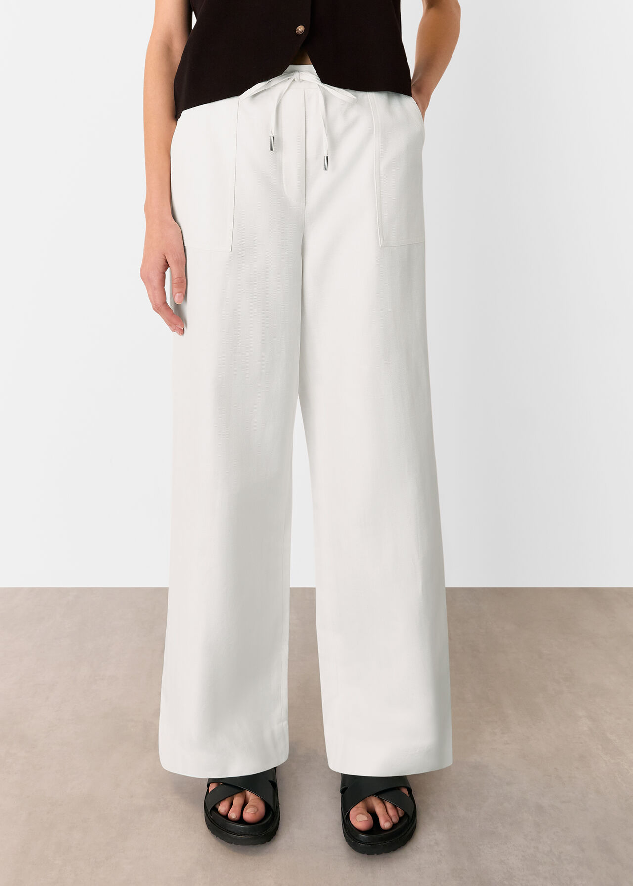 A pair of white linen blend trousers with a wide-leg cut, drawstring waist, and large front pockets, worn by a person standing against a plain background.