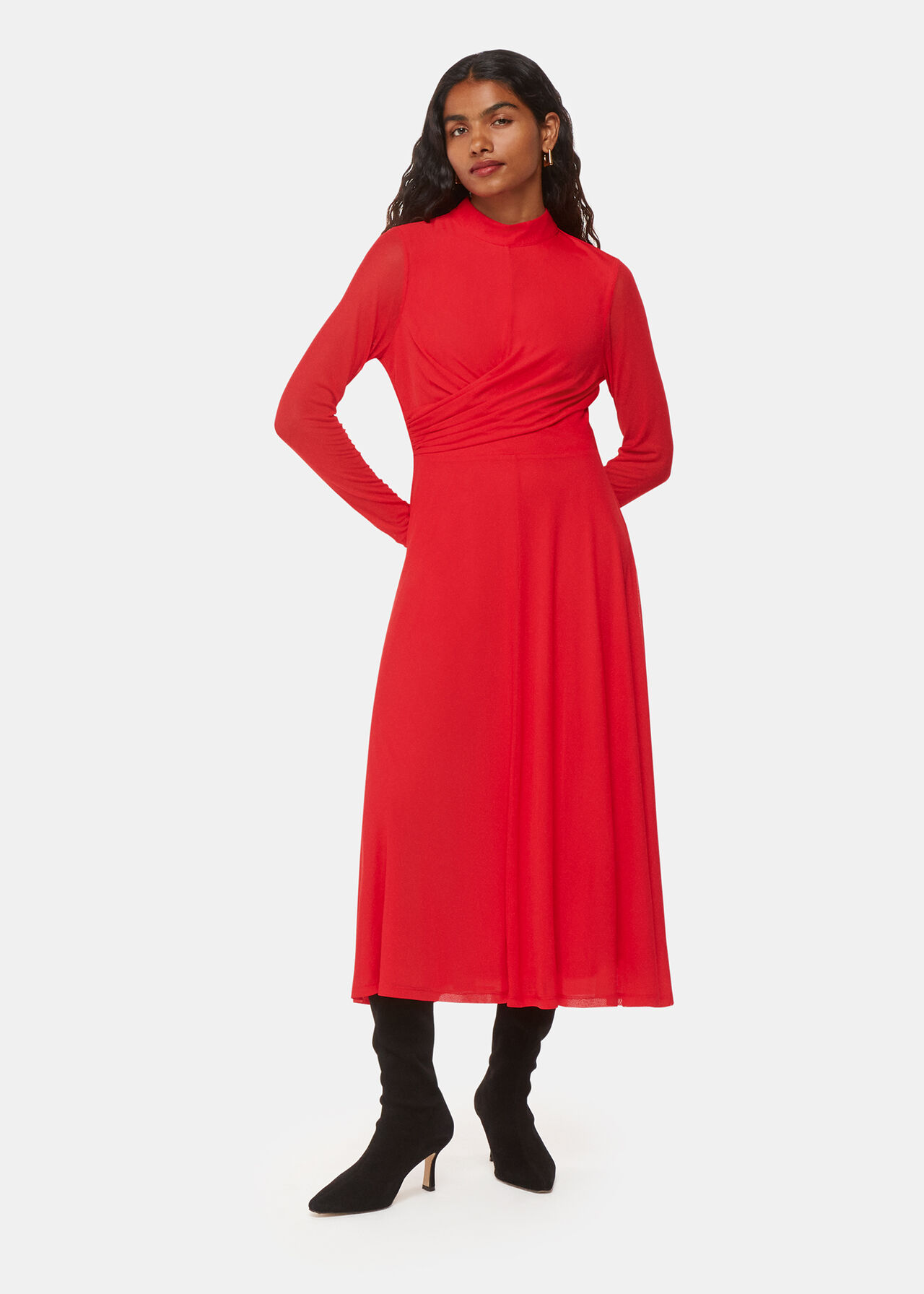 A woman wearing a vibrant red midi dress with long semi-sheer mesh sleeves, draped bodice, and pleated hem, paired with black high-heeled boots.