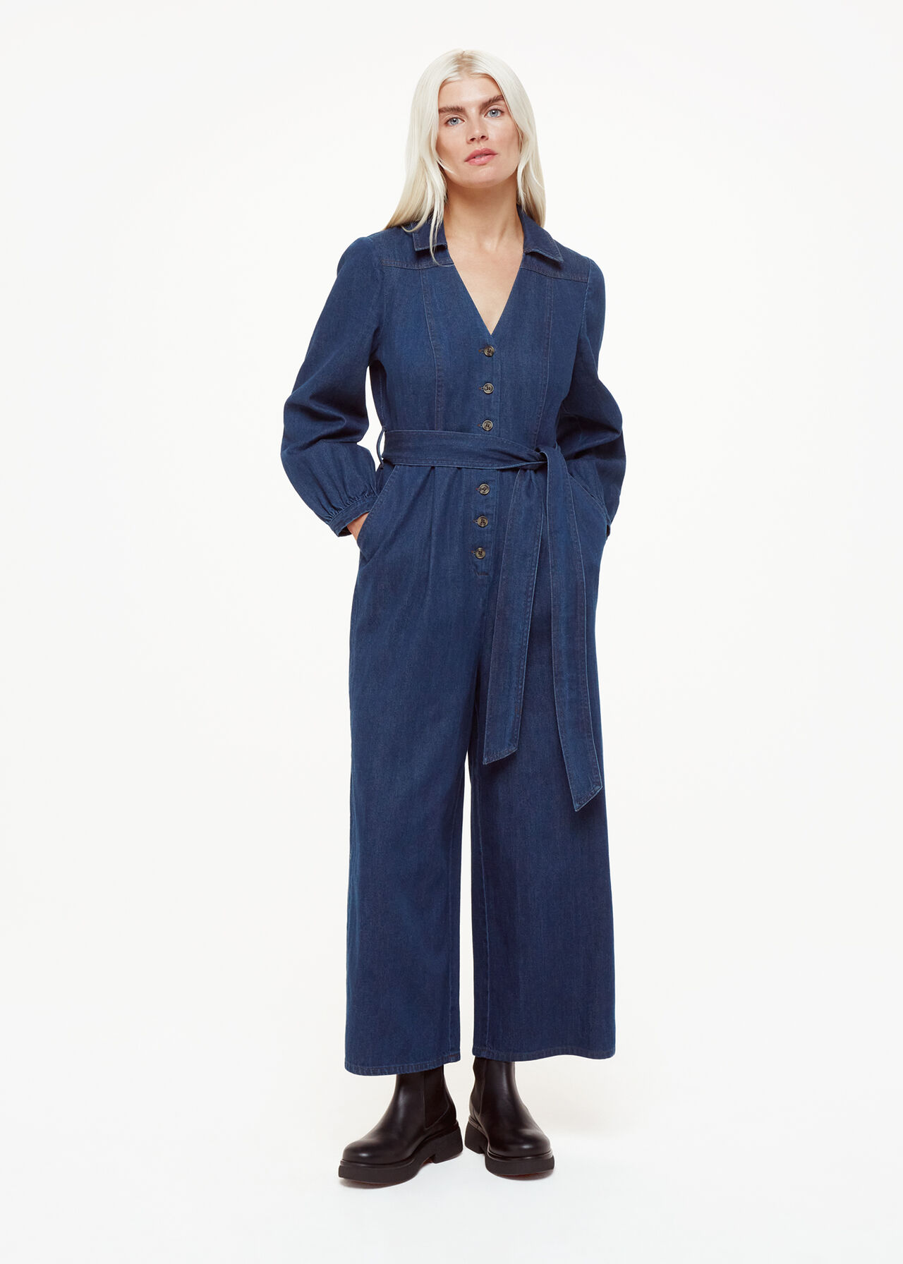 A woman wearing a blue denim belted jumpsuit with long sleeves, a button-down front, and wide-leg pants, paired with black boots.