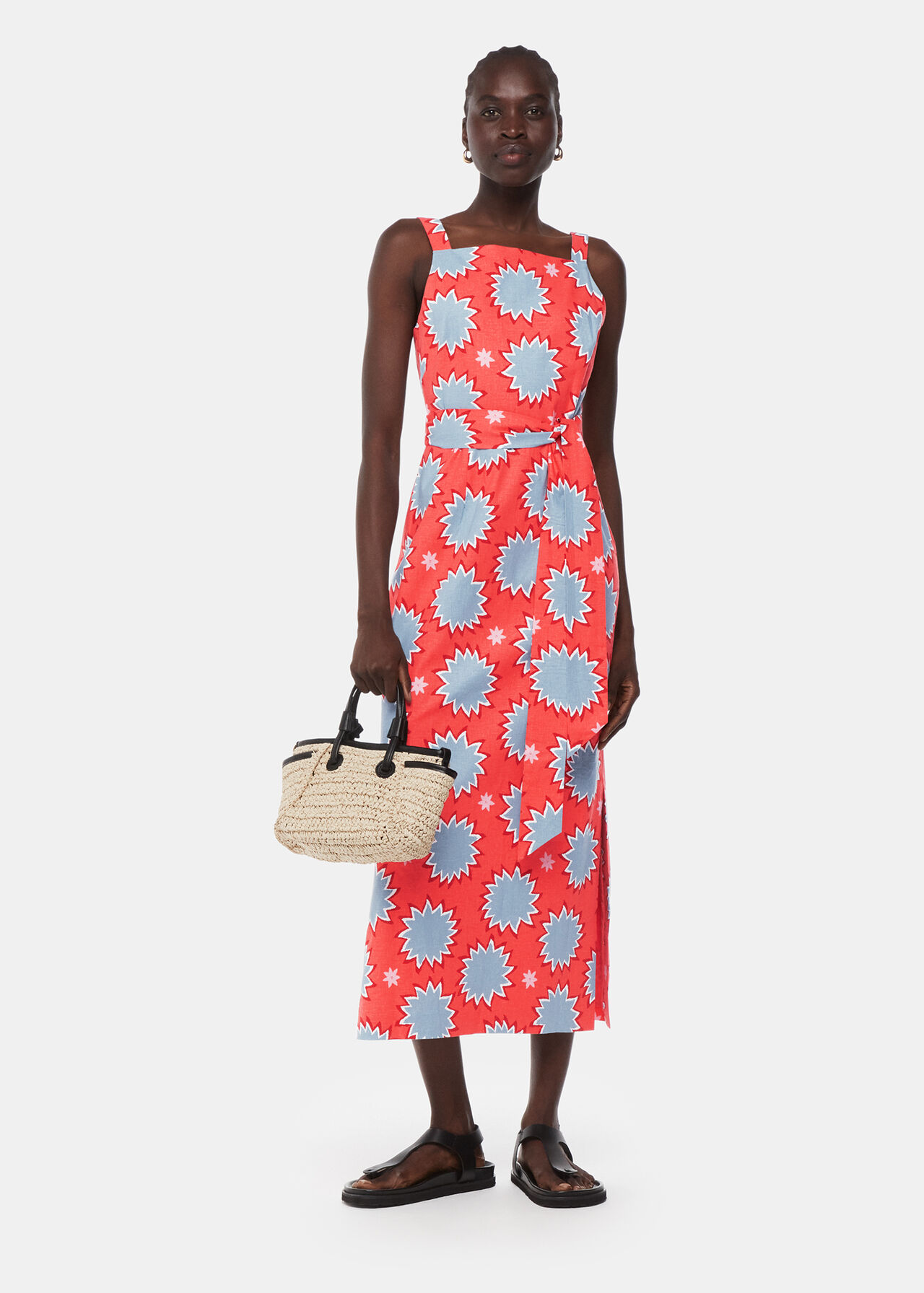 A woman wearing a coral-colored linen blend dress with a sunburst pattern in light blue and white, featuring thick straps and a waist belt, paired with black sandals and a straw handbag.