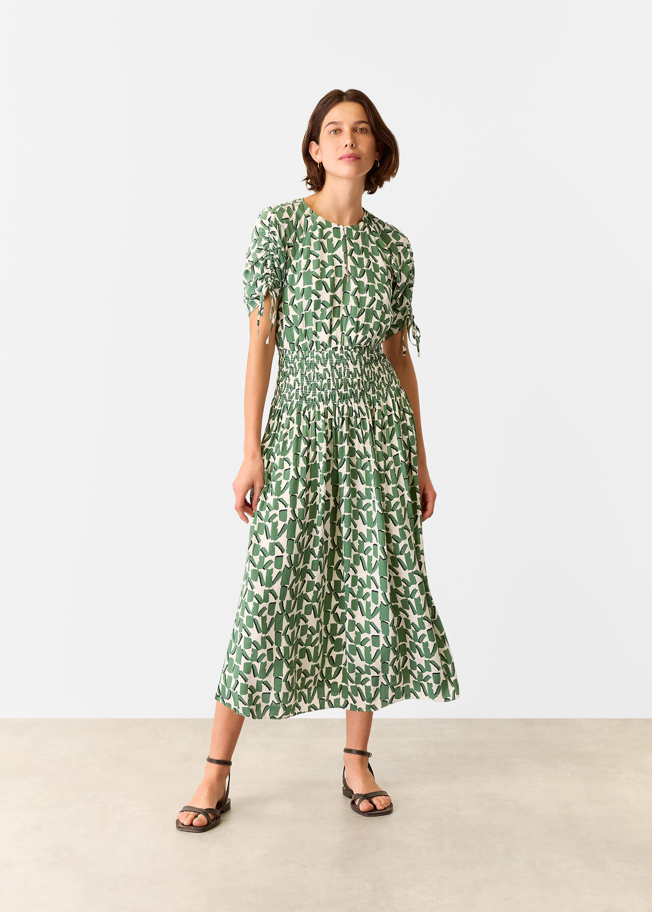 A woman wearing an ivory midi dress with a layered green abstract square pattern, round neckline, front keyhole, ruched short sleeves with drawstrings, shirred waist, and midi length.