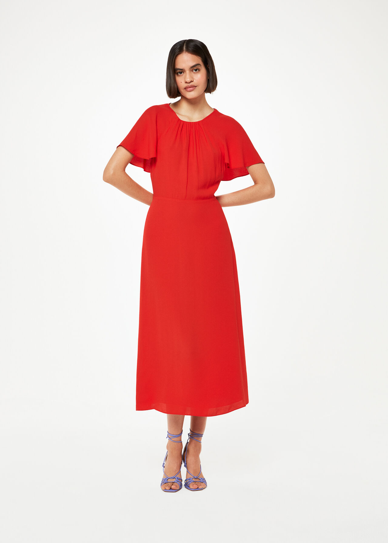A woman wearing a red midi dress with cape sleeves and smocking detail, standing against a plain background.