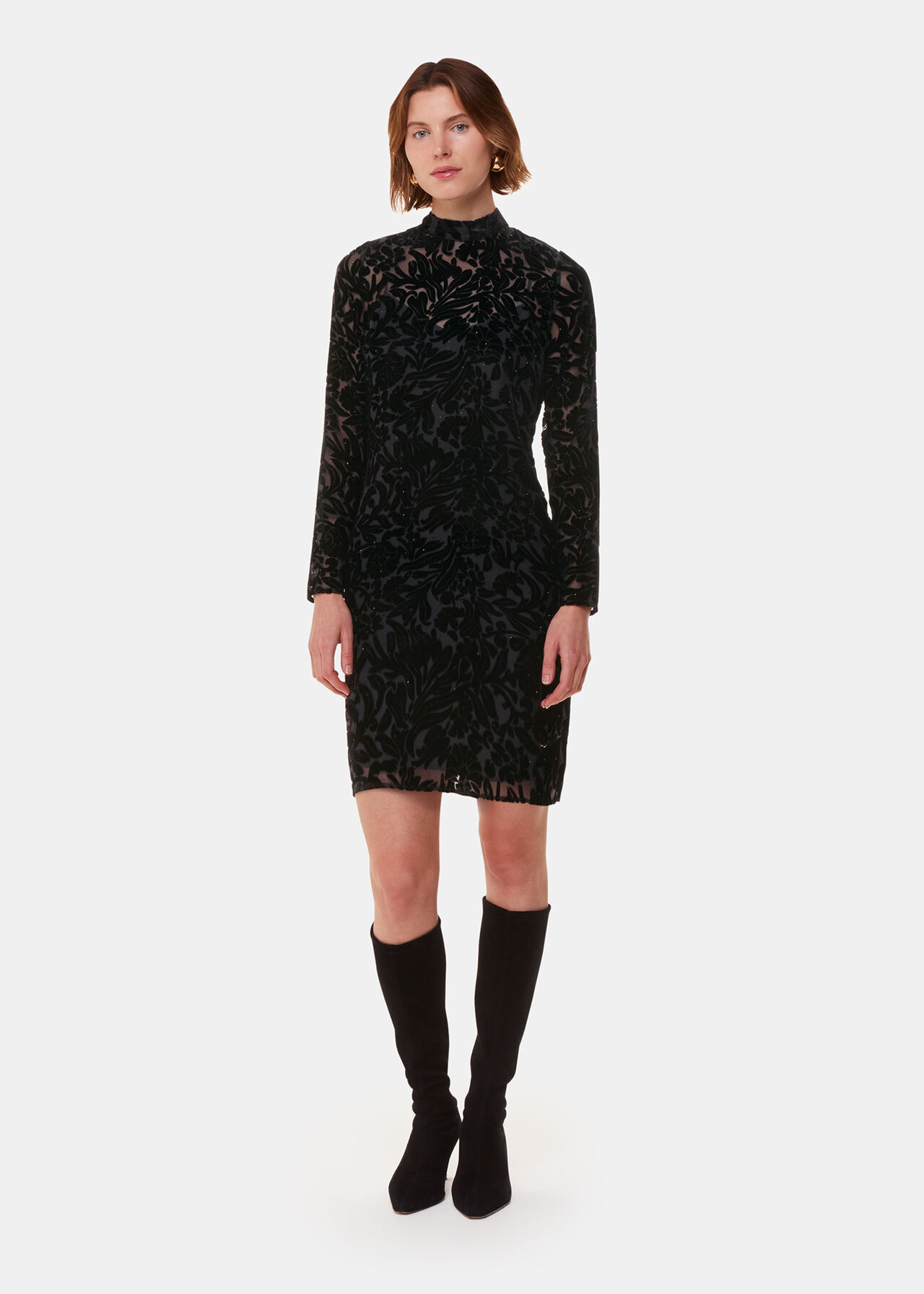A woman wearing a black floral velvet dress with long sleeves and a high neck, standing against a plain background.