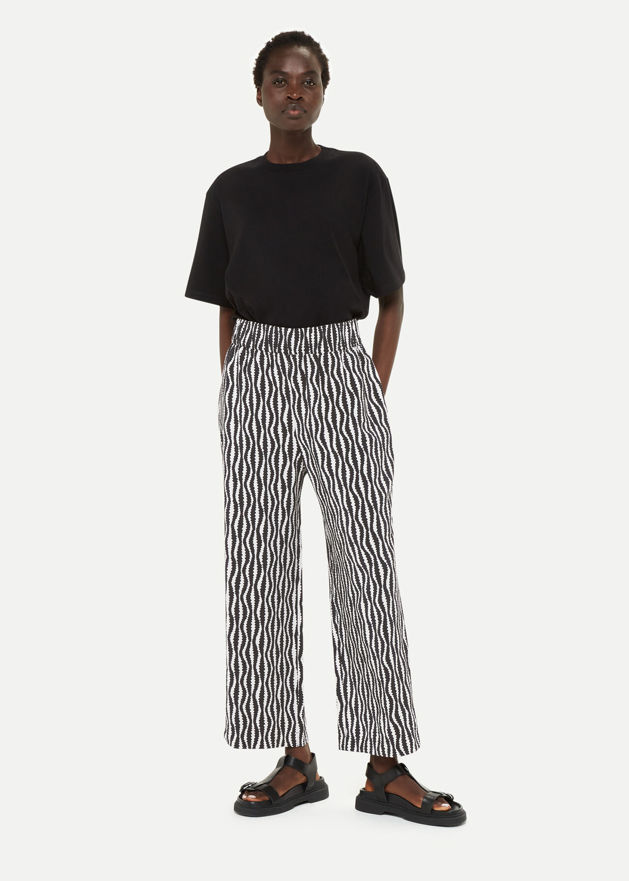 A woman wearing black cropped trousers with a white and black geometric pattern, paired with a black top and black sandals.