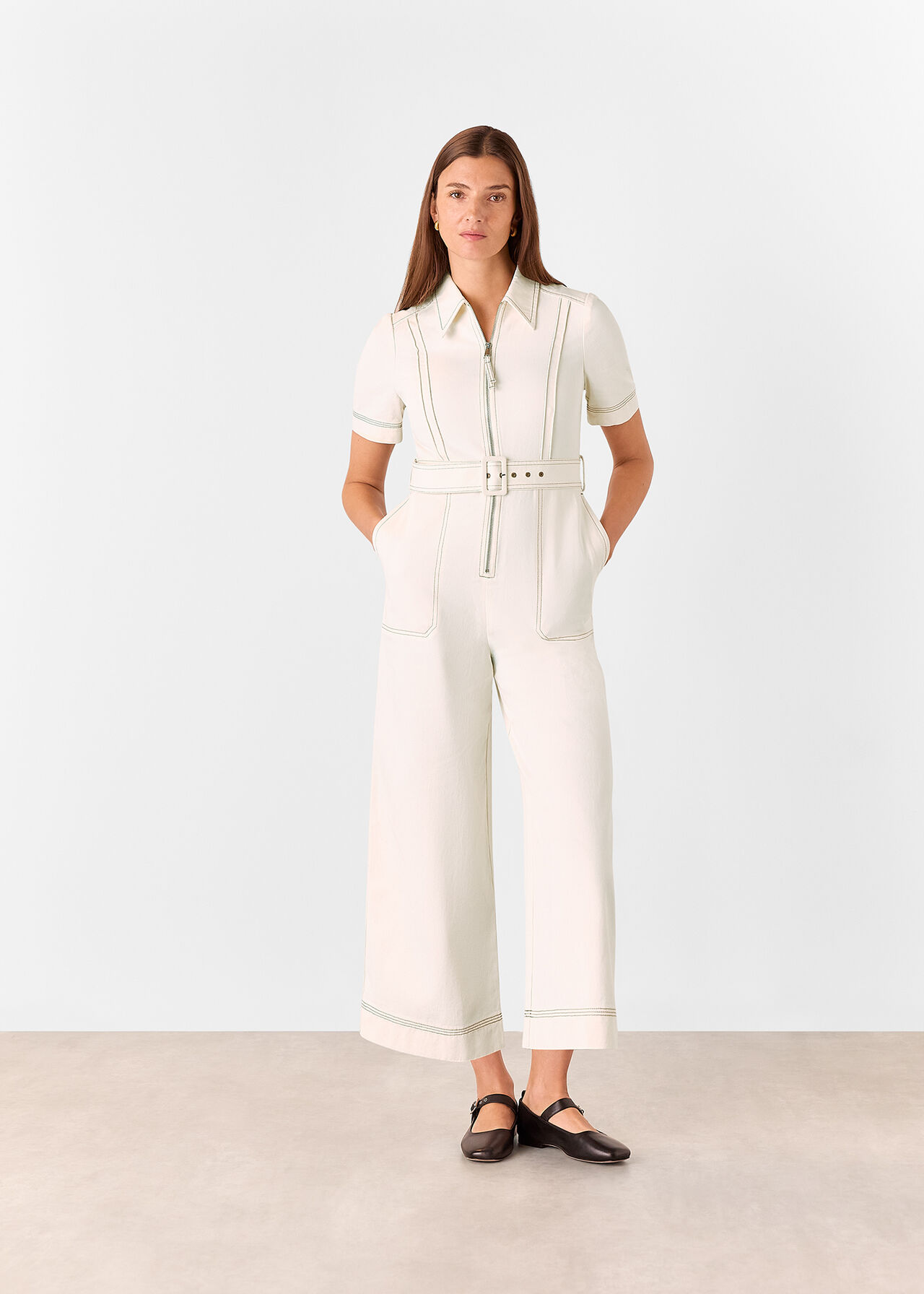 A woman wearing a white denim jumpsuit with a belted waist, short sleeves, a collared neckline, and large side pockets.