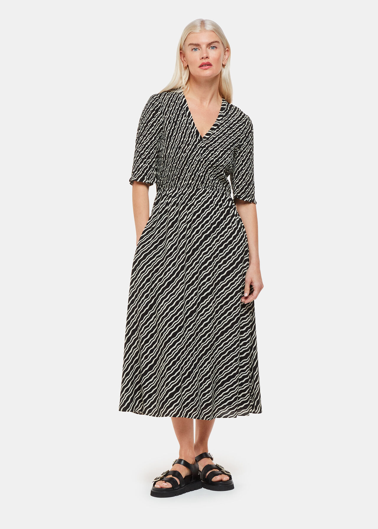 A woman wearing a black and white diagonal ripple print dress with a shirred bodice, elbow-length sleeves, and a centre back zip fastening.