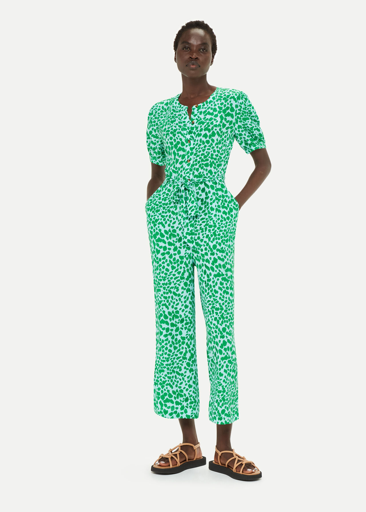 A woman wearing a green and white leopard print jumpsuit with short puffed sleeves, a round neckline with a small slit, and a waist tie, made from lightweight fabric.