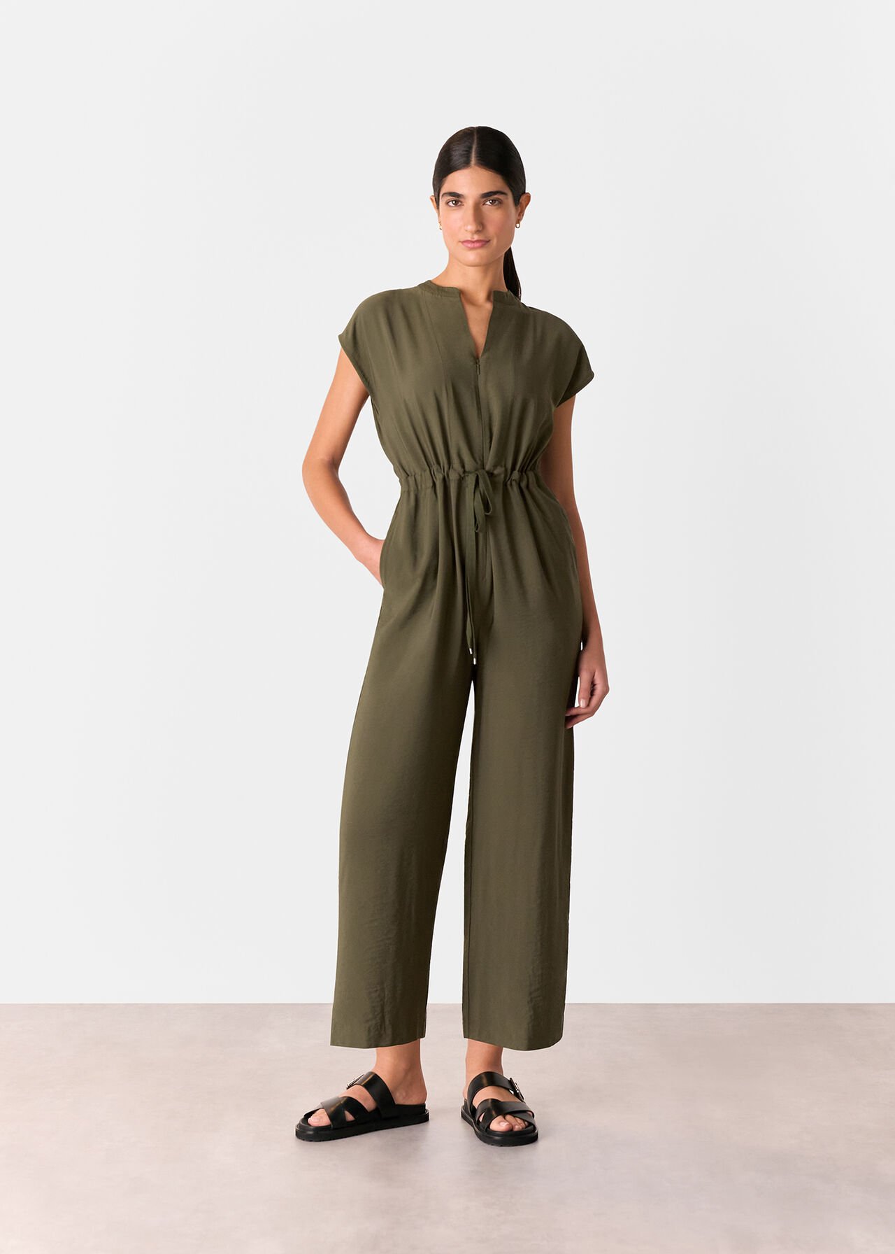A woman wearing a khaki sleeveless jumpsuit with a V-neckline and drawstring waist, standing against a plain background.
