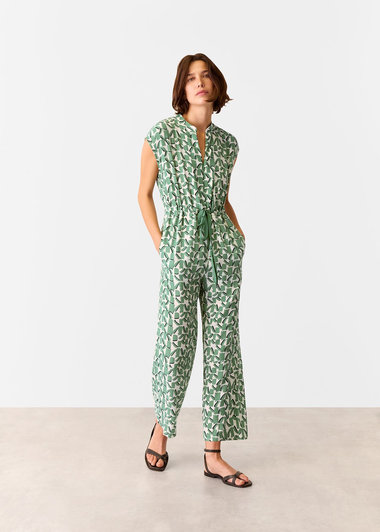 A woman wearing an ivory jumpsuit with green and white abstract square print, short sleeves, drawstring waist, and relaxed-fit legs.