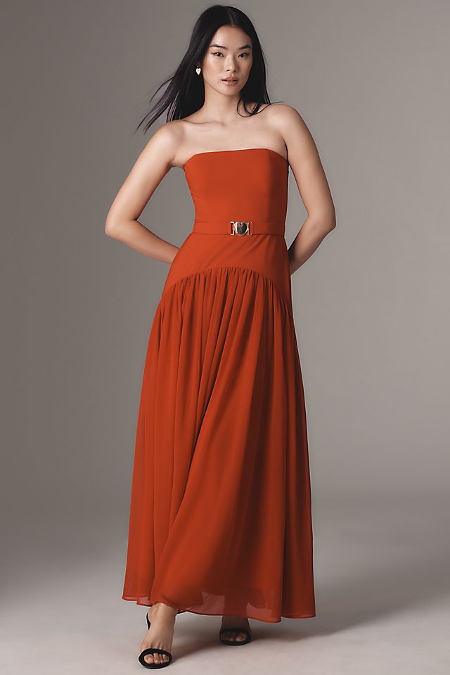 A woman wearing a strapless rust-colored maxi dress with a fitted bodice, a matching belt, and a flowing pleated skirt.