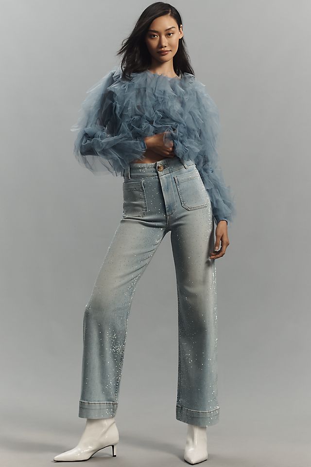 A woman wearing light-wash, high-rise, cropped wide-leg jeans with a sparkly finish, paired with a sheer, ruffled blue top and white boots.