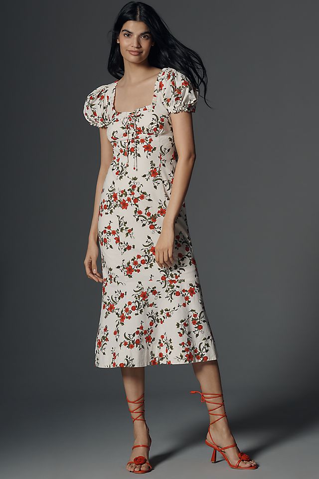 A woman wearing a cream-colored midi dress with red and black floral print, puffed short sleeves, sweetheart neckline, and a flowing skirt, paired with red strappy heels.