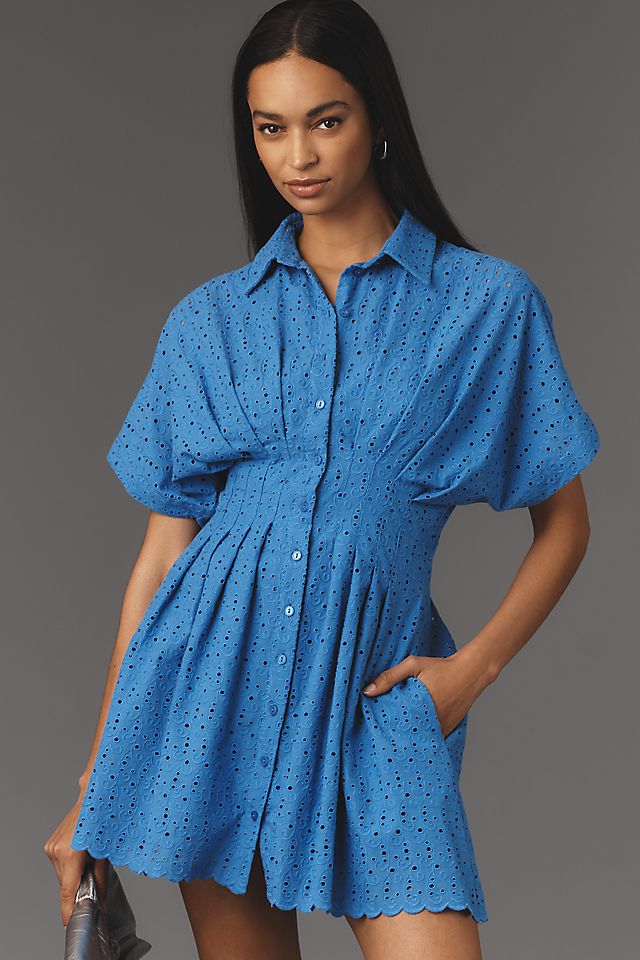 A woman wearing a bright blue eyelet shirt dress with a classic collar, short puffed sleeves, and a pleated skirt, with her right hand in her pocket.