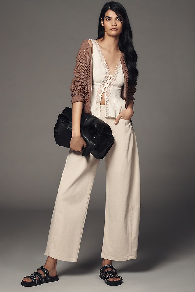 A woman wearing beige mid-rise barrel jeans, a light-colored top, a brown cardigan, and black sandals, holding a black bag.