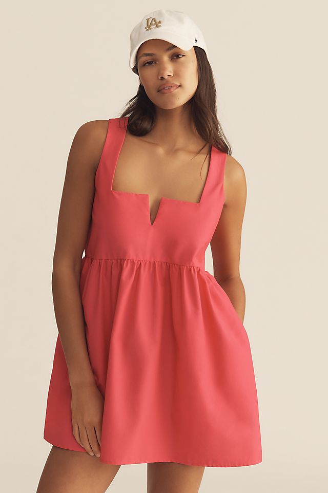 A woman wearing a sleeveless pink mini dress with a notch neckline and a gathered skirt, paired with a white baseball cap.