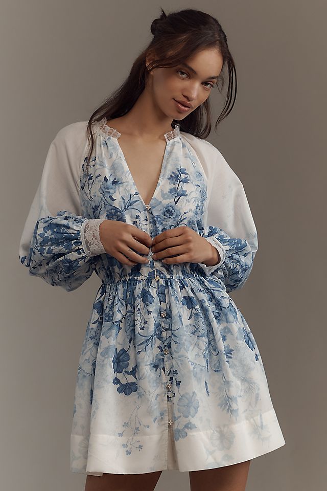 A woman wearing a white mini dress with blue floral patterns, V-neckline, and long sleeves with lace cuffs.