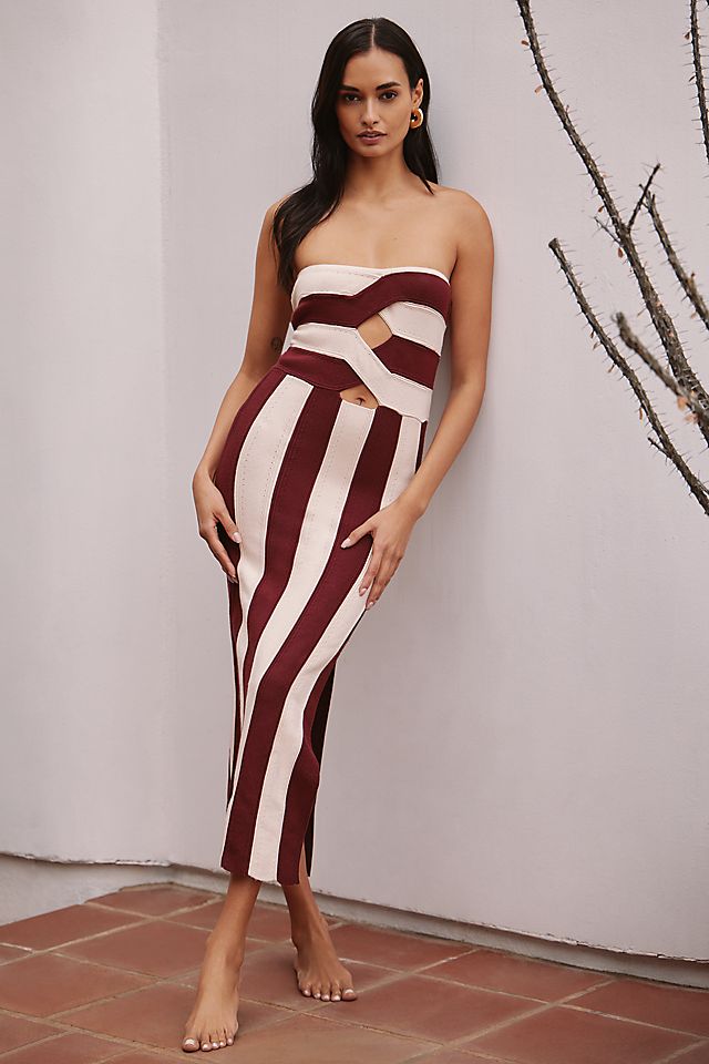 A woman wearing a strapless, cream and deep red striped knit midi dress with front cutouts, standing barefoot against a plain background.