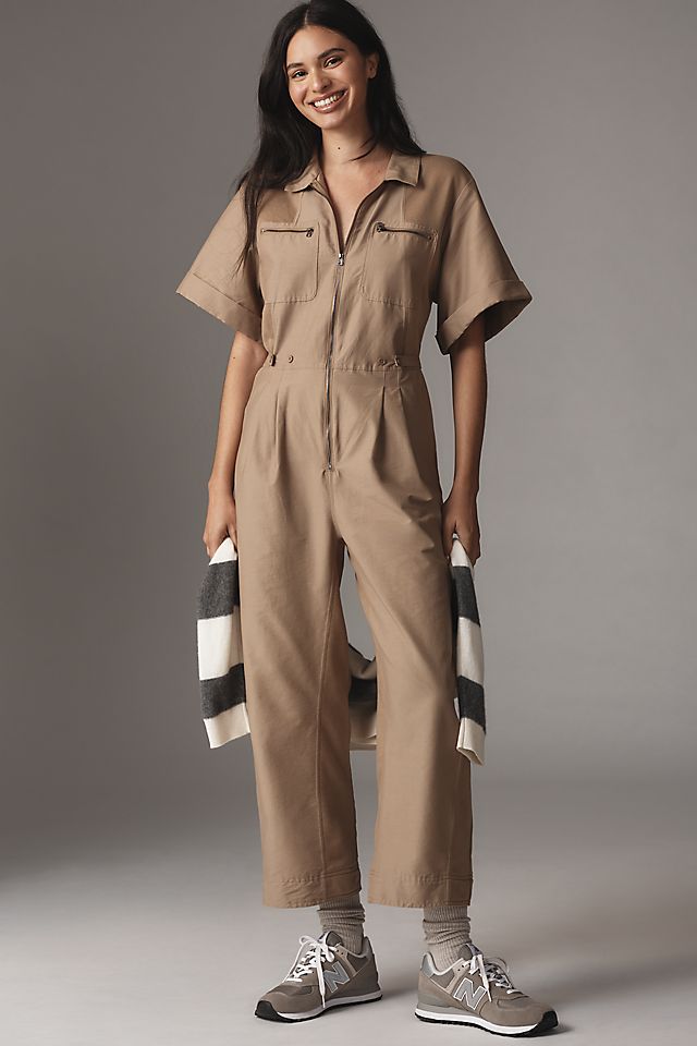 A woman wearing a beige short-sleeve utility jumpsuit with a front zipper and chest pockets, paired with sneakers and holding a striped scarf.