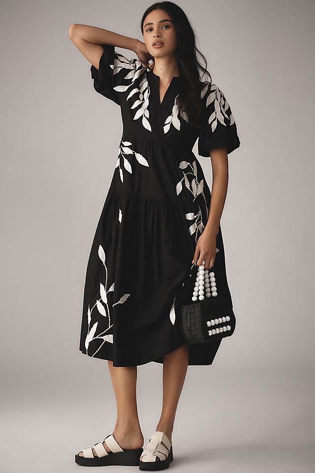 A woman wearing a black midi dress with short sleeves and white embroidery appliqué details, holding a black handbag with white accents, and wearing white sandals.
