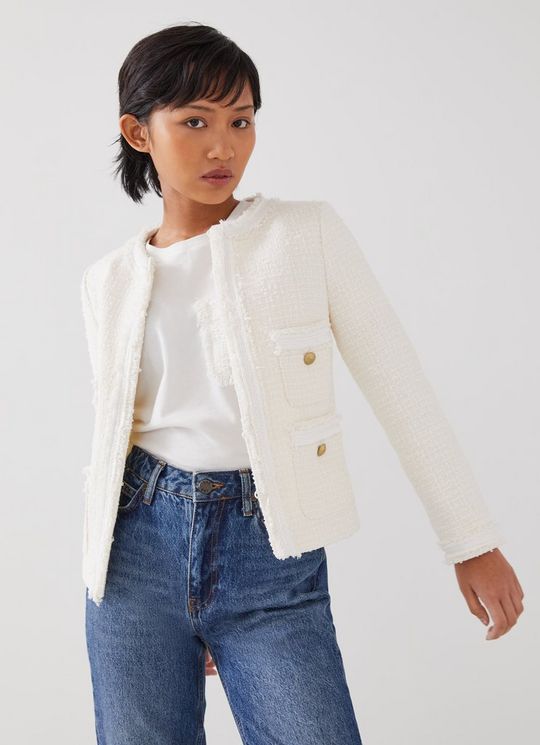 A woman wearing a collarless ivory tweed jacket with black trim, patch pockets, and gold buttons, paired with a white top and blue jeans.