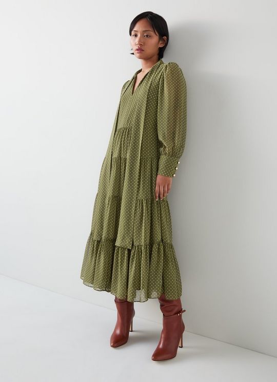 A woman wearing an olive and birch polka dot tiered dress with a tie-neck and full-length sleeves, paired with brown high-heeled boots.