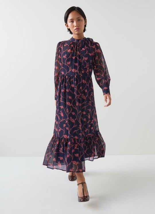 A woman wearing a navy blue and bright pink floral print maxi dress with a tie neck, long sleeves, and ruffled hem.