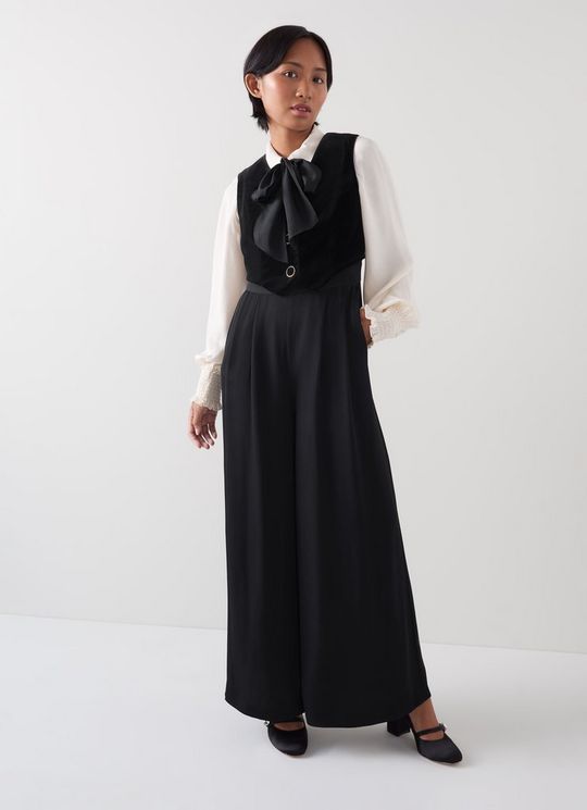 A woman wearing a black waistcoat culotte jumpsuit with a sleeveless waistcoat top, V-neck, wide-leg trousers, and a back zip, styled with black shoes.