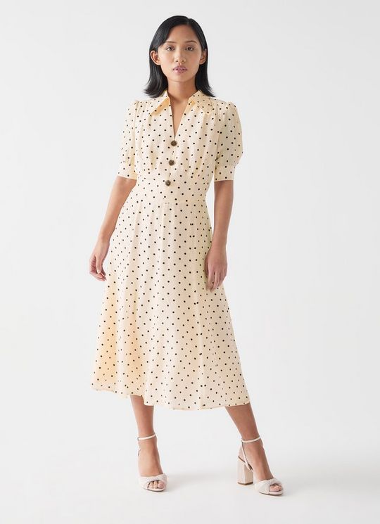 A woman wearing a cream-colored, polka dot midi dress with a collared V-neckline, black buttons, and puffed short sleeves.