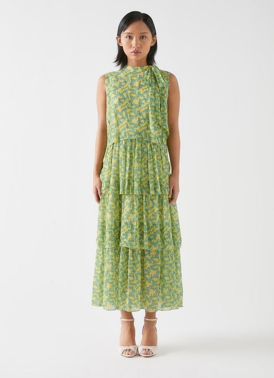 A woman wearing a sleeveless, tiered, blossom print dress in yellow and green, with a tie neck and picot edging, standing against a plain background.