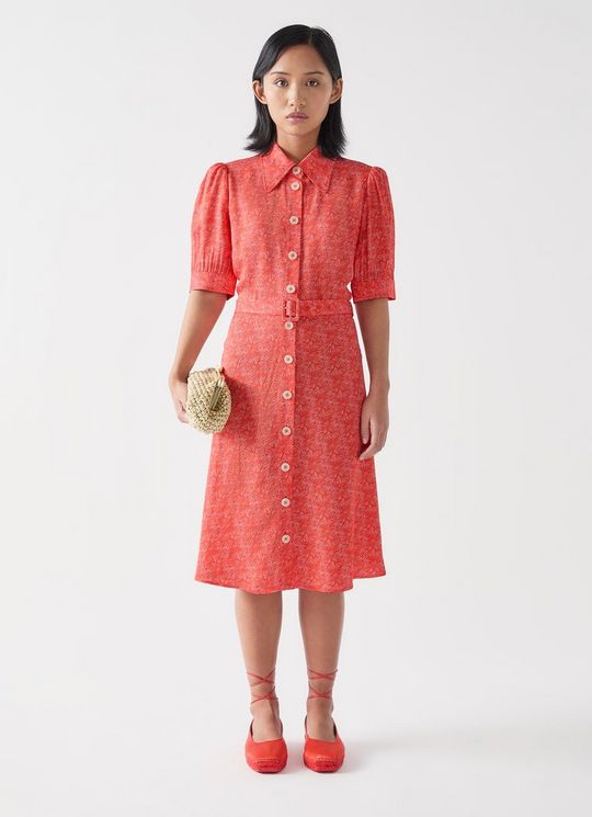 A woman wearing a pink and red swirl print shirt dress with full-length sleeves, a button-down front, and a belt, paired with red shoes and holding a small woven clutch.