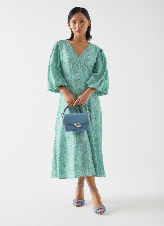 A woman wearing a green floral jacquard midi dress with statement sleeves, V-neckline, and a fit and flare silhouette, holding a blue handbag and wearing blue heels.