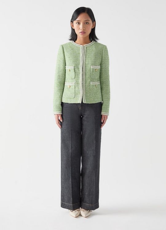 A woman wearing a green collarless tweed jacket with fringe trims and crest metal buttons, paired with dark wide-leg trousers.